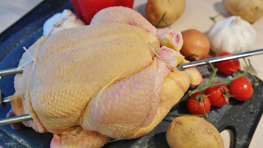 Picture of a raw chicken ready for grilling with potatoes, tomatoes, onions, and a head of garlic nearby.