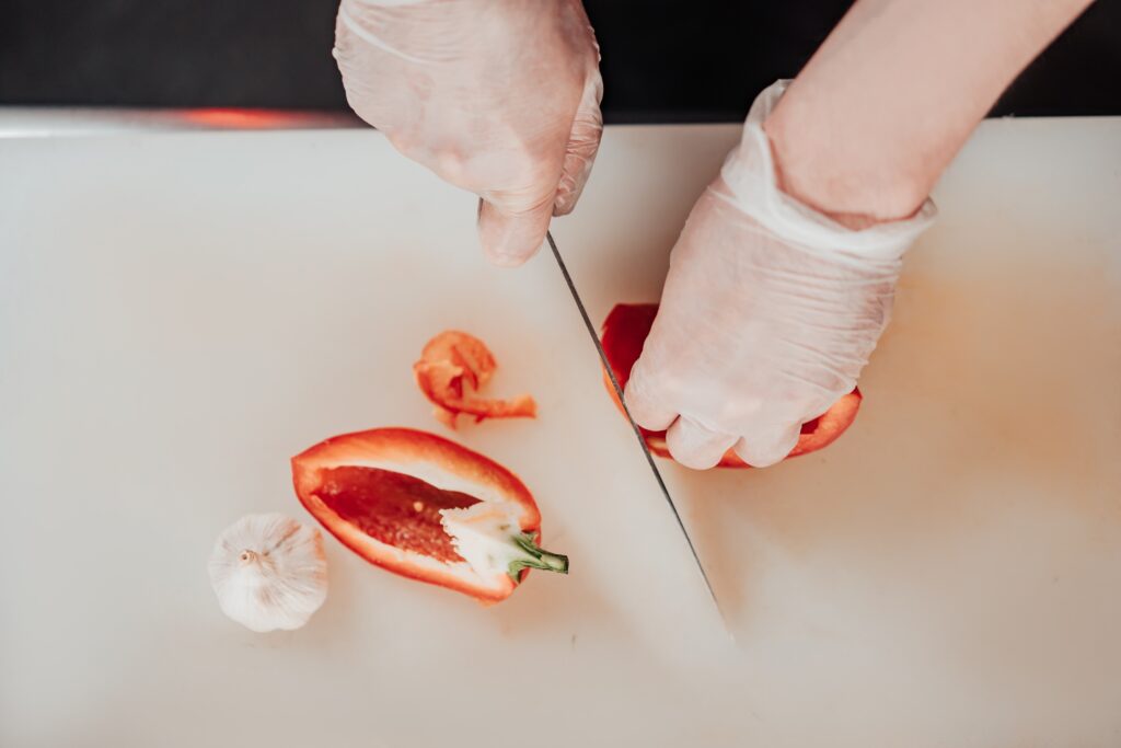 A pair of hands wearing gloves cutting up a red chili on a white chopping board. The gloves protect me from the capsaicin and demonstrate food safety