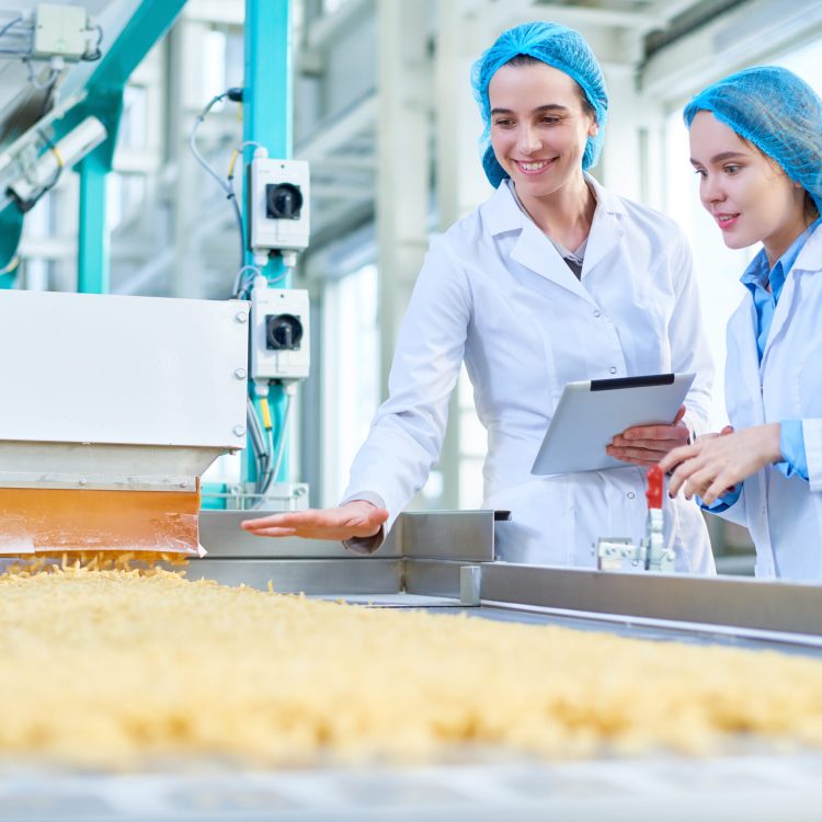 Waist up portrait of  two young female workers wearing lab coats standing by  conveyor line with macaroni  in clean production workshop, copy space